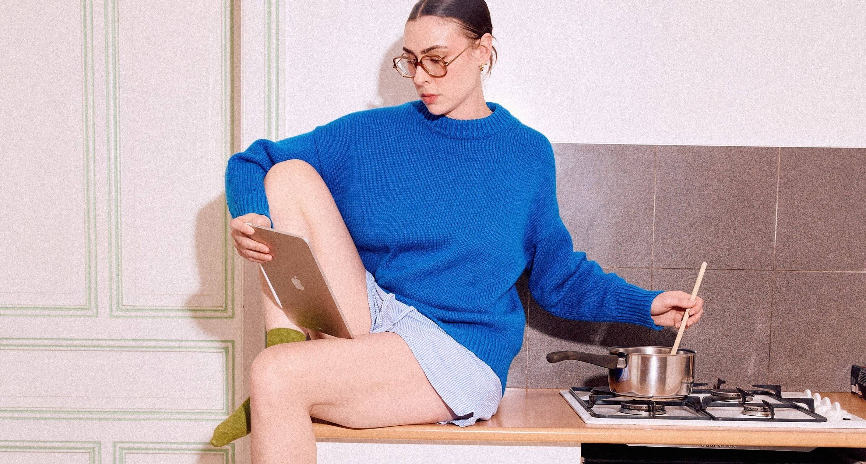 femme en pull bleu assise sur le plan de travail d'une cuisine avec une tablette dans une main et une cuillère dans l'autre