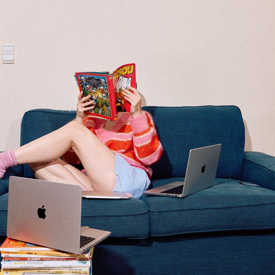 femme sur un canapé lisant un magazine avec des ordinateurs autour d'elle